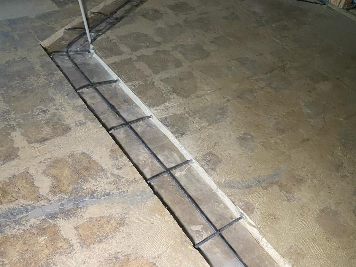 A long trench cut and patched across a bare concrete floor in a gutted building, with a pipe stub rising at one end
