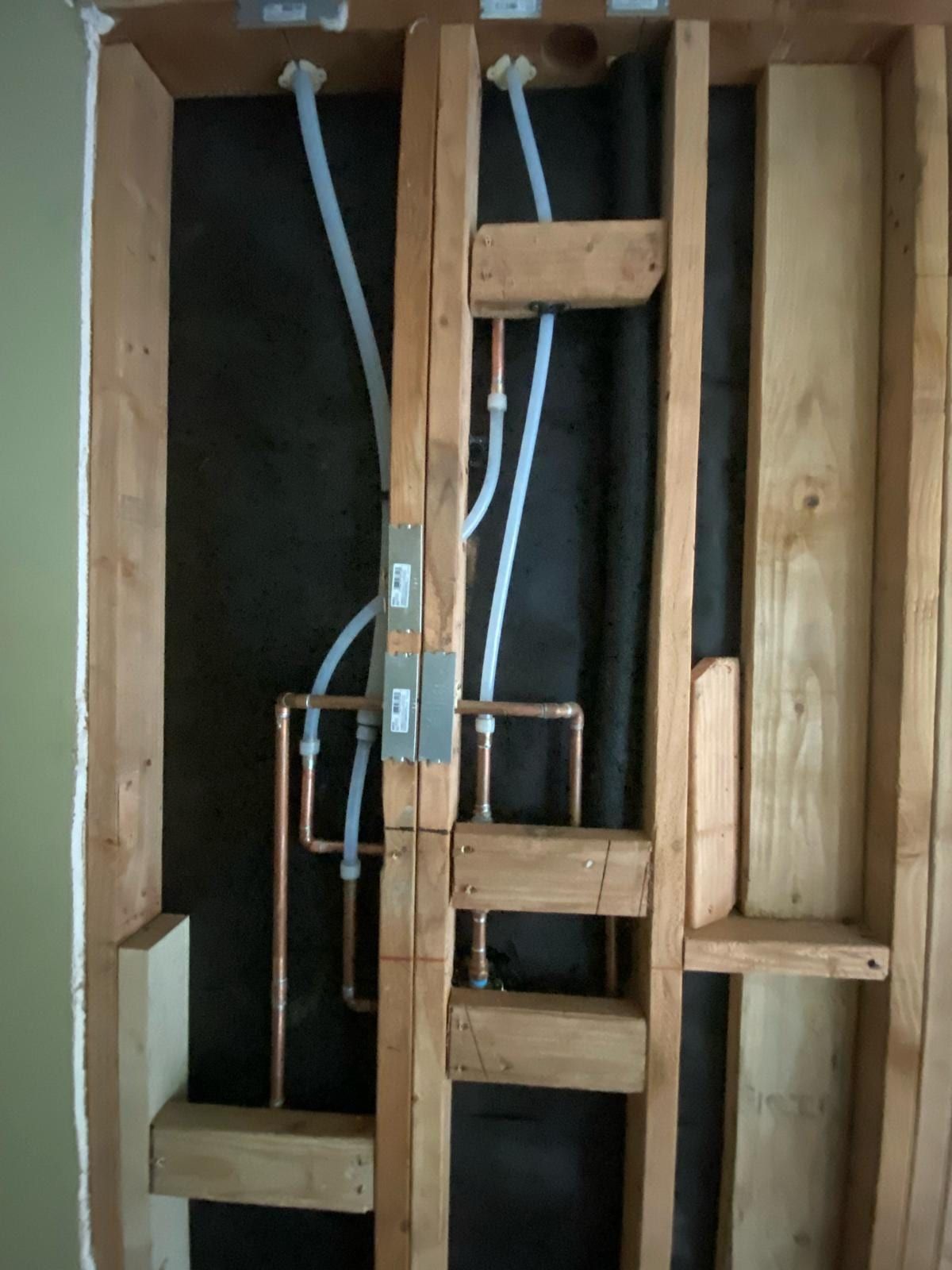 Open wall framing where new PEX lines drop from the top plate and tie into copper pipe, with metal nail plates on the studs