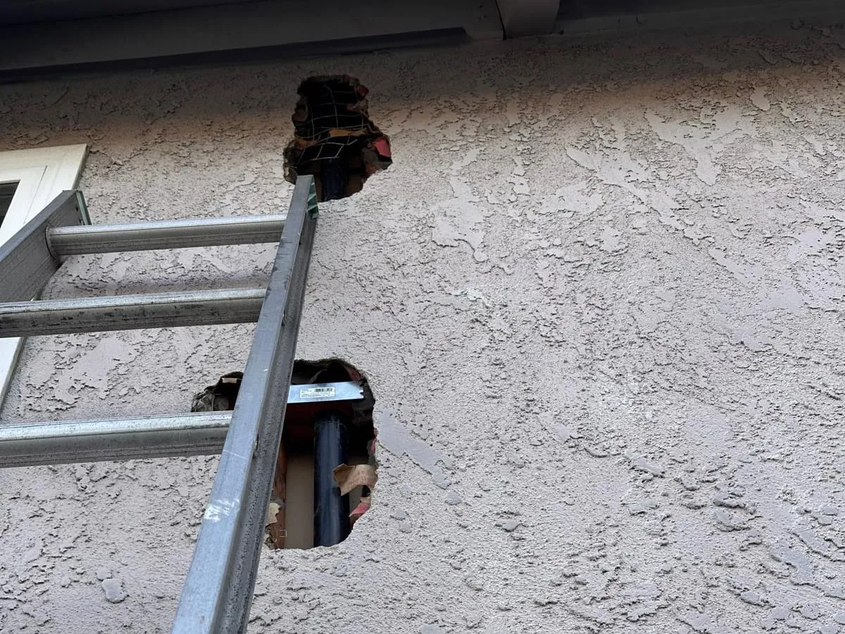Two openings cut in a stucco exterior wall beside an aluminum ladder, with pipe visible inside the wall