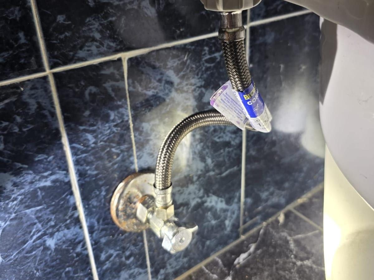 A new braided supply line, its tag still on, running from a chrome angle stop to the bottom of a toilet tank, against dark marble-look tile