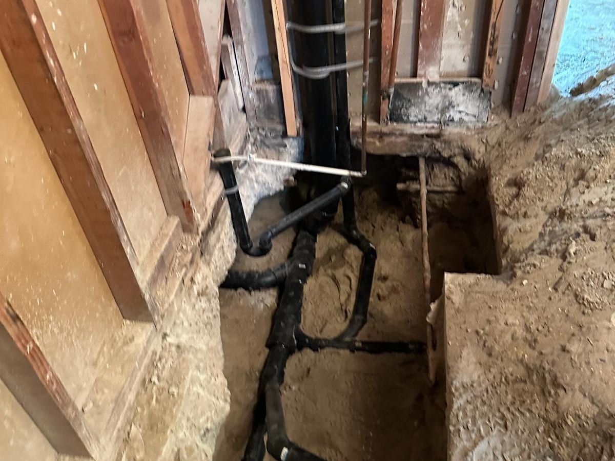 A trench dug along the bottom of an opened wall, with black ABS drain pipe and fittings laid in the dirt
